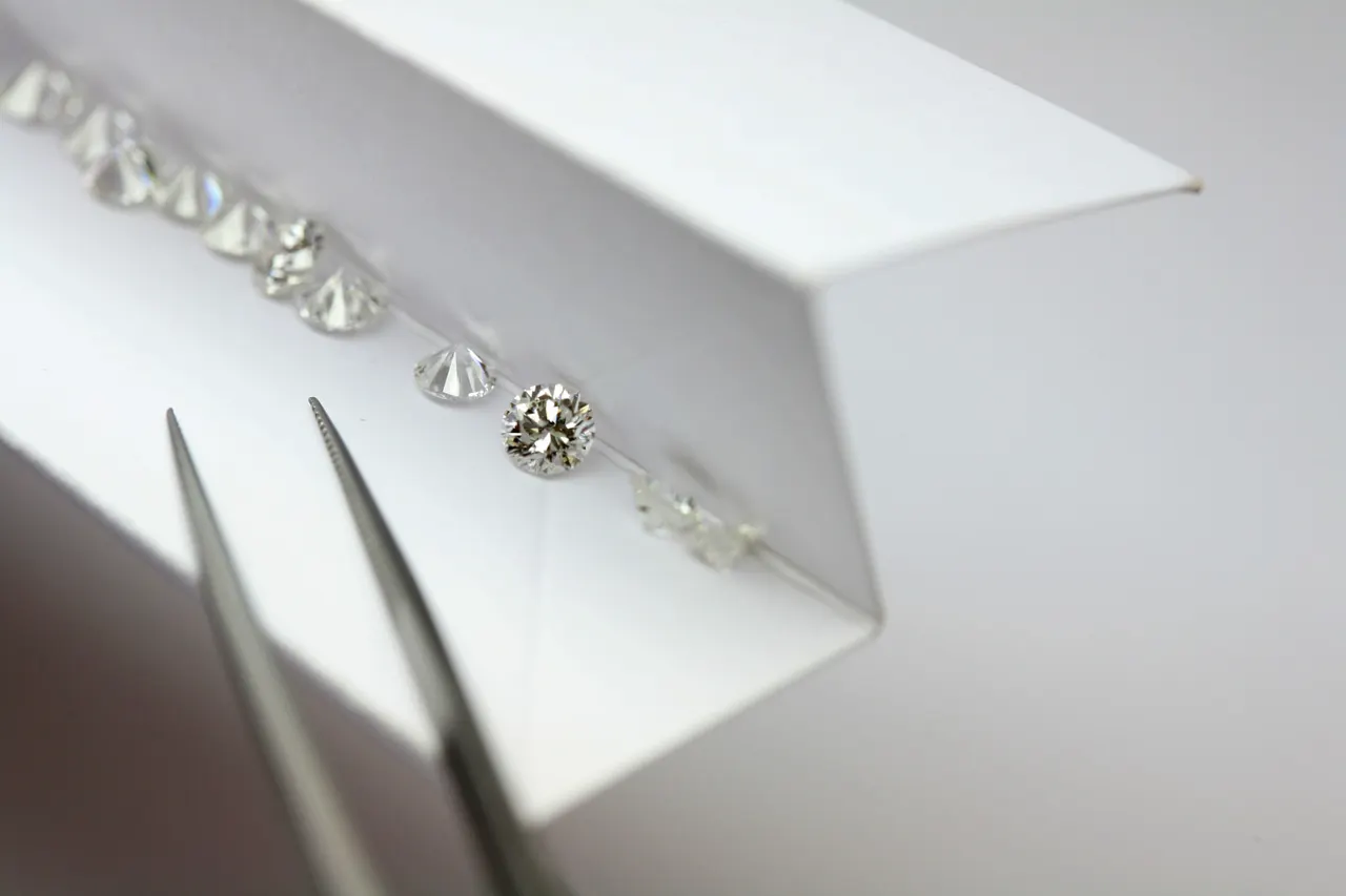 Round brilliant diamonds lined up in a white parcel box beside precision tweezers.
